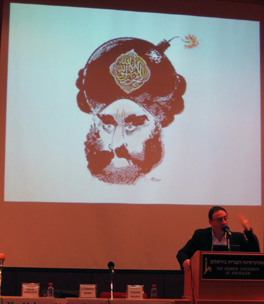 הקריקטוריסט מישל קישקה על רקע קריקטורה של הנביא מוחמד