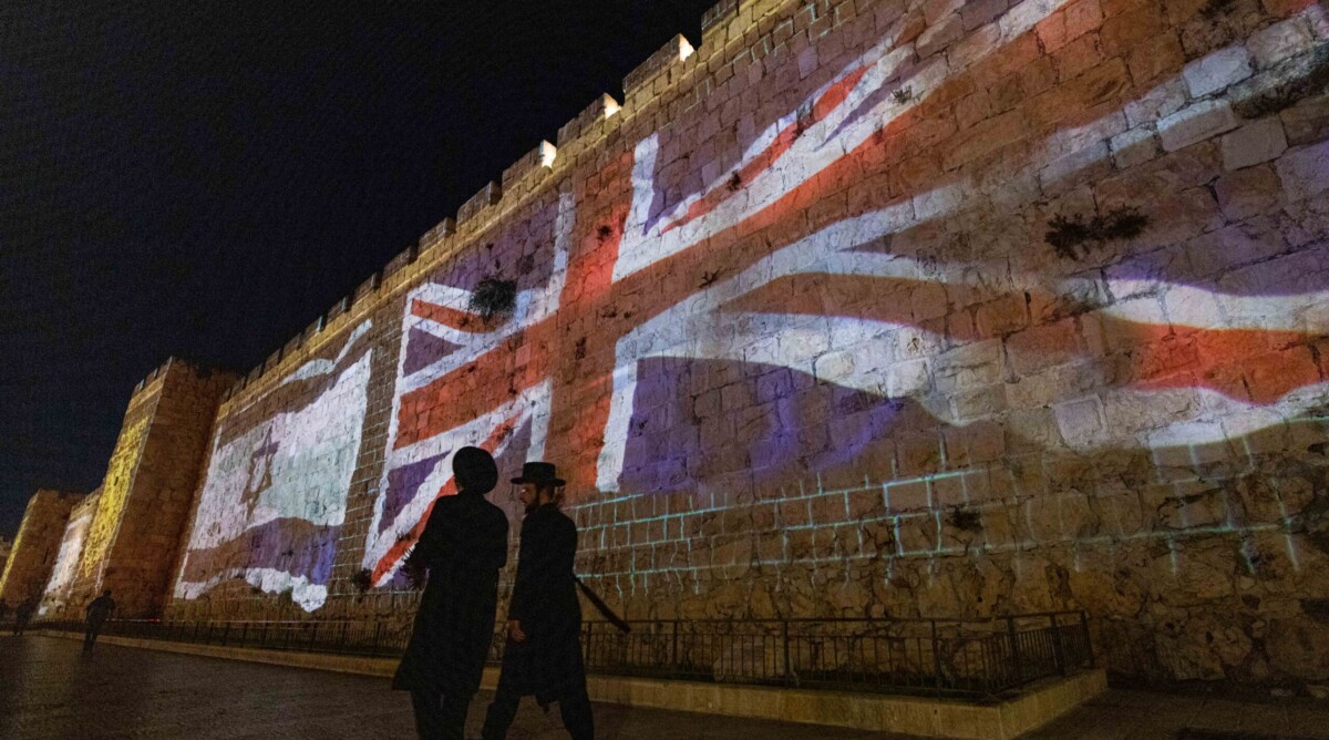 צמד ישראלים חולפים על פני חומת העיר העתיקה בירושלים, שעליה מוקרנים דגלי בריטניה וישראל לאות הזדהות עם האבל על מות המלכה אליזבת. 8.9.2022 (צילום: יונתן זינדל)