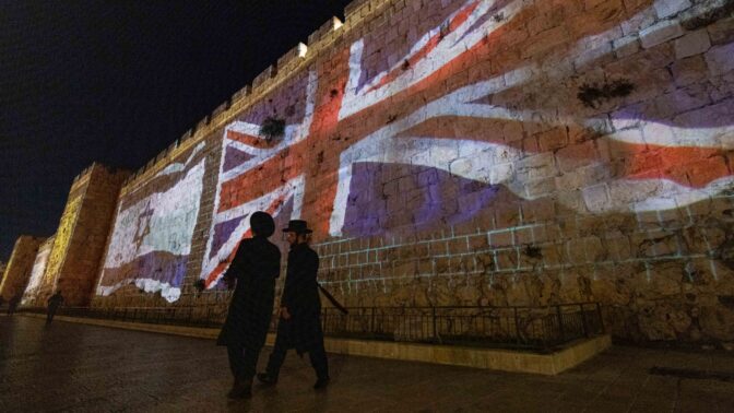 צמד ישראלים חולפים על פני חומת העיר העתיקה בירושלים, שעליה מוקרנים דגלי בריטניה וישראל לאות הזדהות עם האבל על מות המלכה אליזבת. 8.9.2022 (צילום: יונתן זינדל)